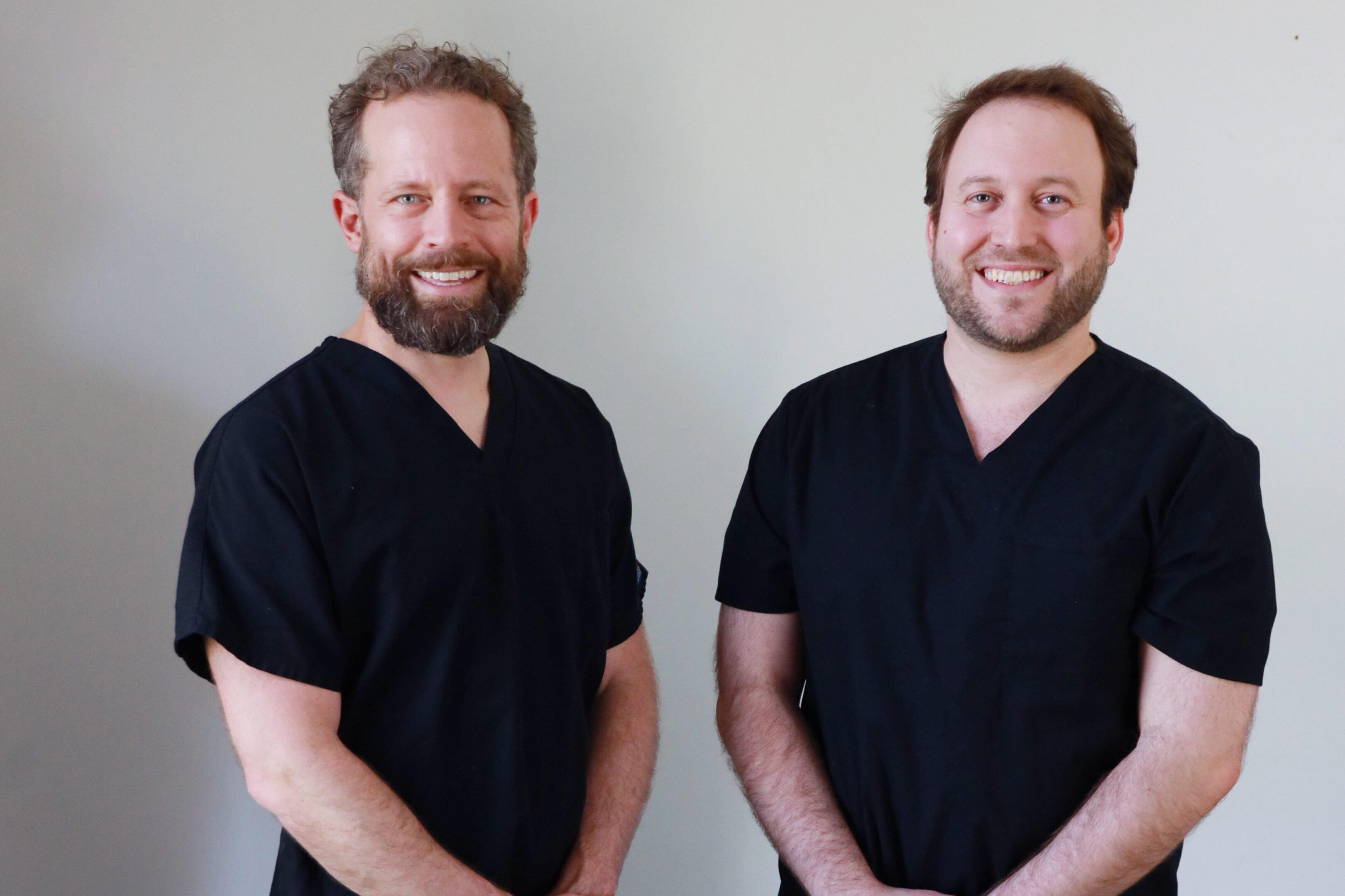 Two men in black medical scrubs smiling against a plain background.