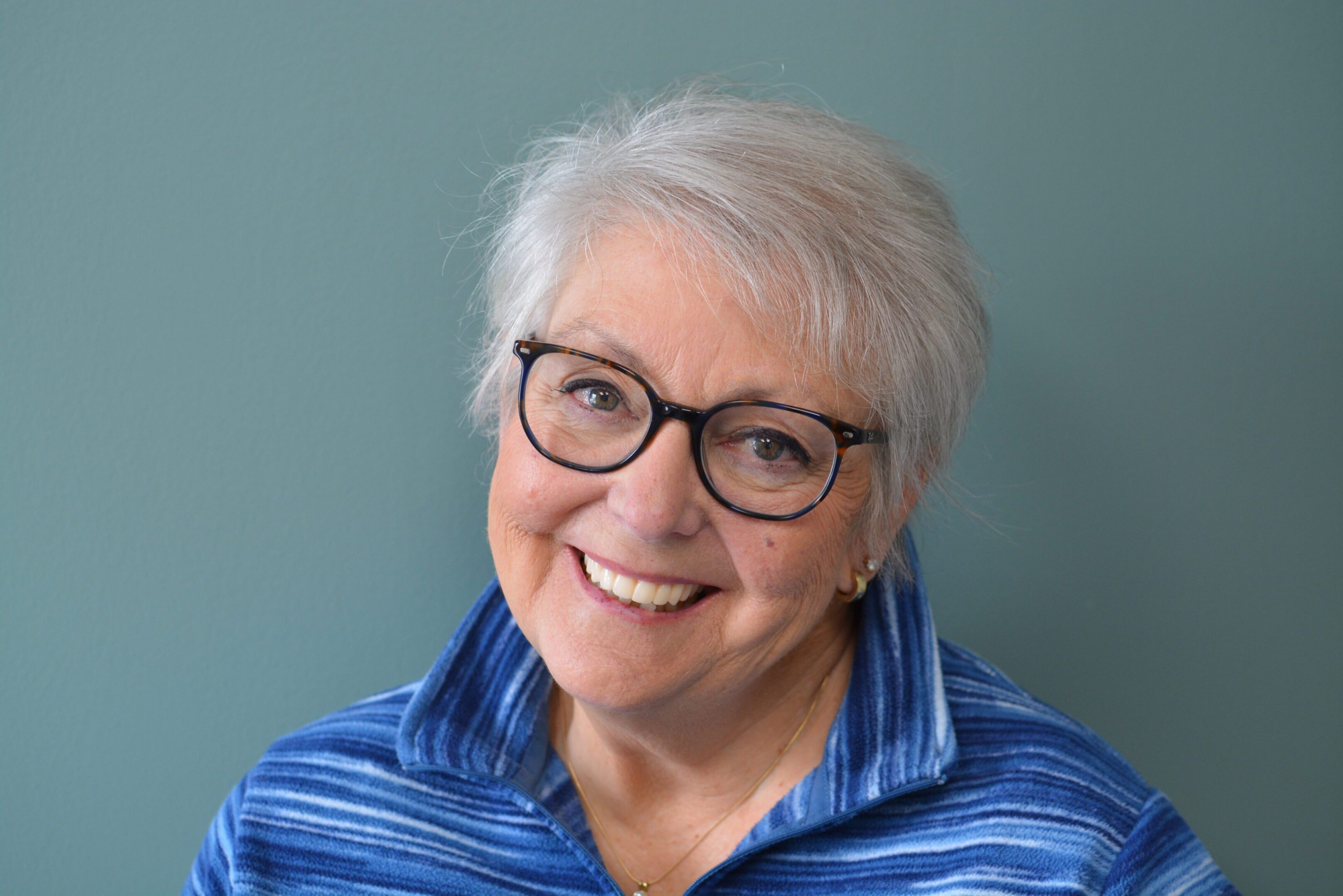 Smiling elderly woman with glasses and gray hair wearing a blue striped shirt.
