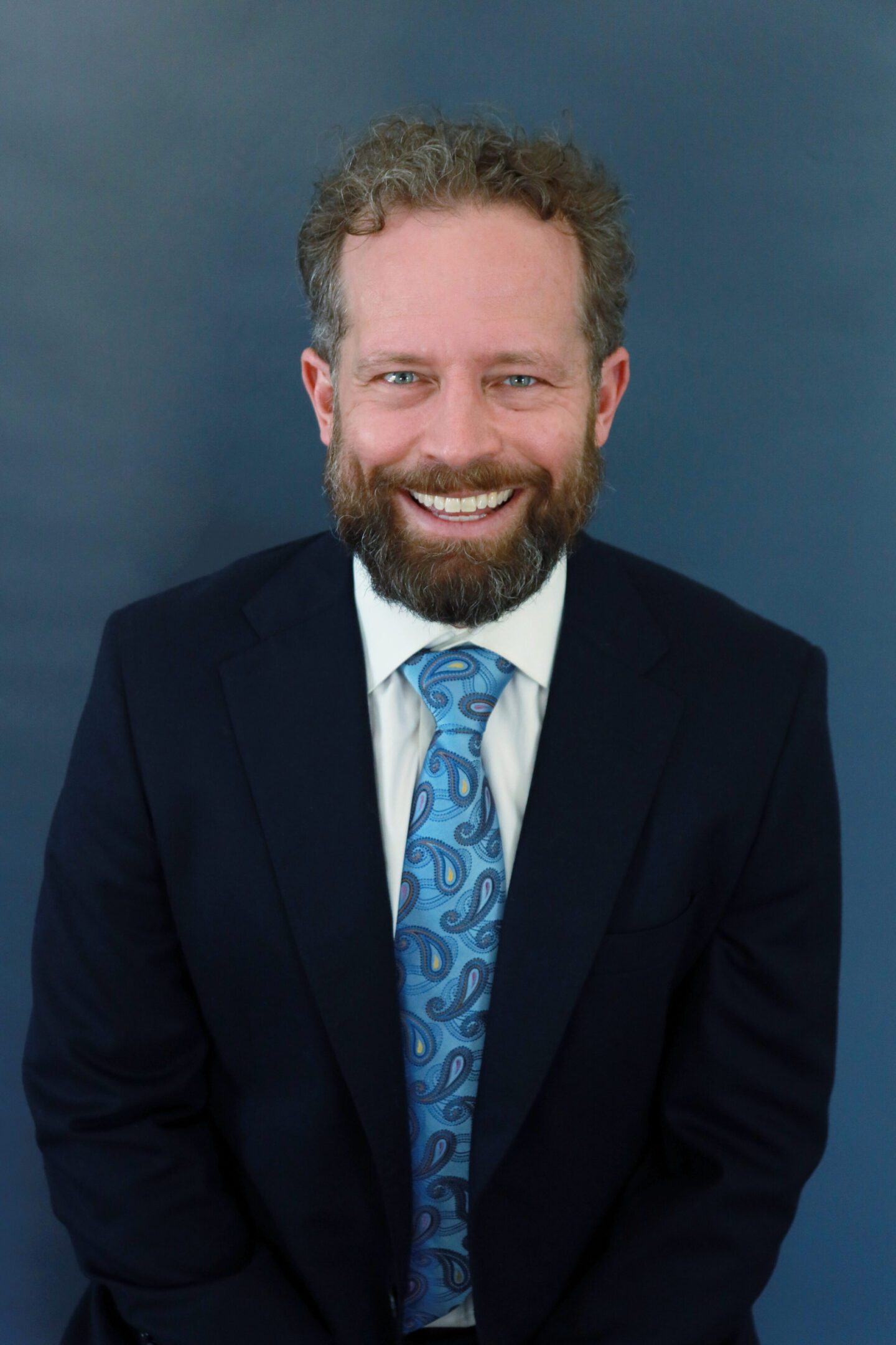 Smiling man in a dark suit and blue patterned tie against a dark background.
